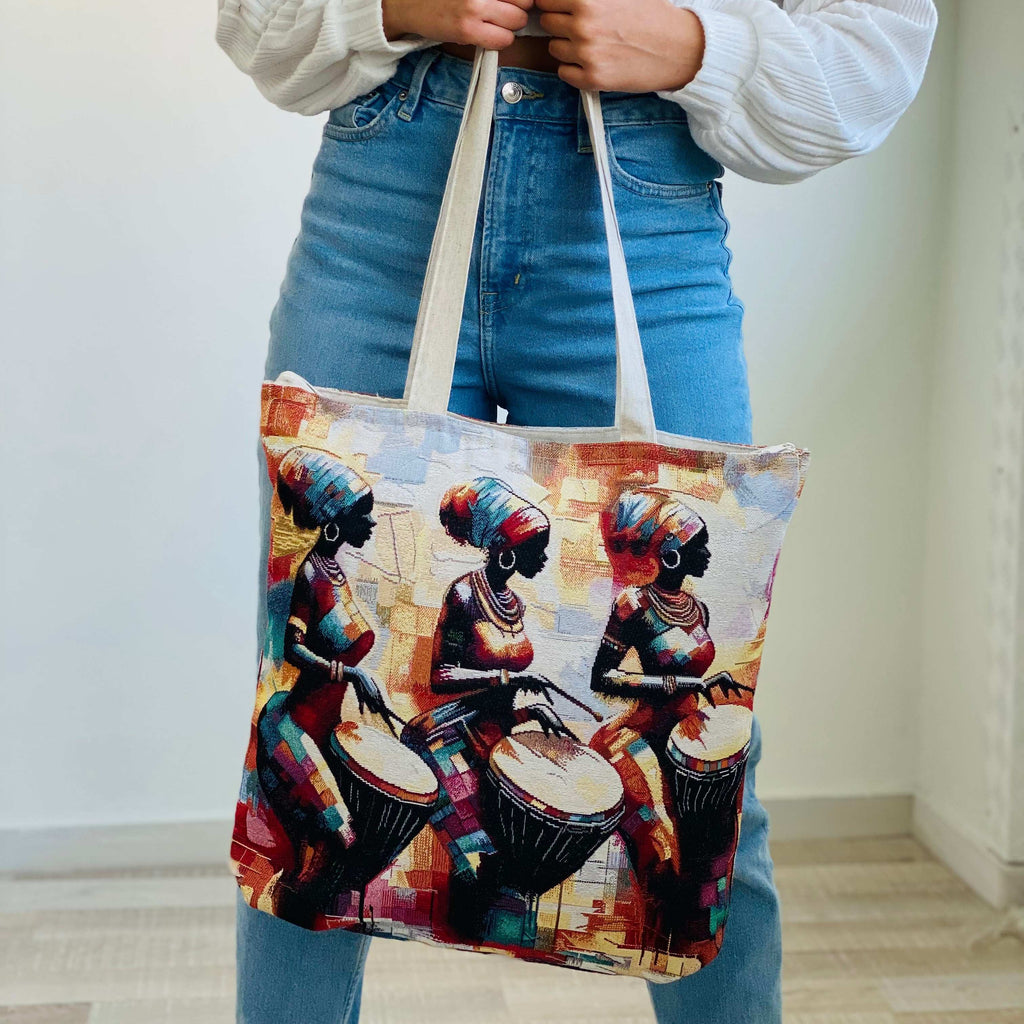Tote bag with an artistic design of people playing drums held by a person wearing blue jeans.